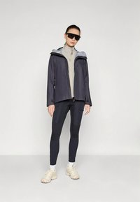 Chaqueta impermeable azul marino con capucha gris, tela ligera, cremallera frontal, combinada con leggings negros y zapatillas beige con suelas texturizadas.