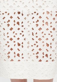 Λευκή δαντελένια φούστα με μοτίβο κοπής λουλουδιών που αφήνει να φαίνεται το δέρμα από κάτω, φτάνοντας πάνω από το γόνατο.