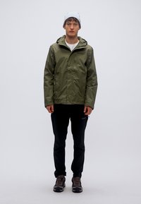 Αδιάβροχο μπουφάν ελιάς με κουκούλα, φερμουάρ μπροστά και ρυθμιζόμενες μανσέτες. Συνδυασμένο με μαύρο παντελόνι και γκρι αθλητικά παπούτσια.