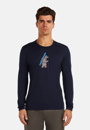 Mann trägt ein marineblaues Langarmshirt mit einer Grafik eines Bären, der Skier und Wanderstöcke trägt, auf der Vorderseite.