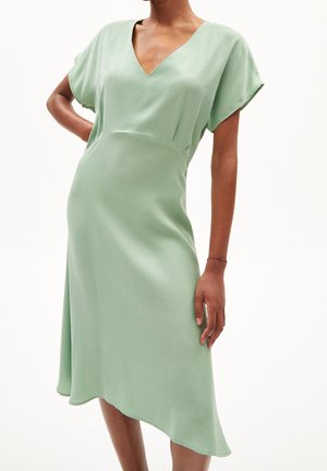 Vestido verde claro, fluido, con escote en V y mangas cortas. El dobladillo es asimétrico, creando una silueta suave y caída.