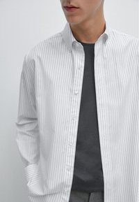 Camicia bianca a maniche lunghe con sottili righe verticali, abbinata a una maglietta grigia antracite sotto, esposta su uno sfondo neutro.