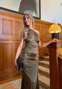 Femme en robe verte satinée à une épaule, debout sur des escaliers moquettés, tenant un sac à main noir, avec des panneaux en bois et une lampe à proximité.