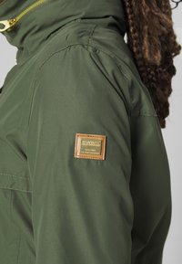 Personne portant une veste vert olive avec un patch rectangulaire beige sur la manche supérieure et une fermeture éclair jaune visible près du col.