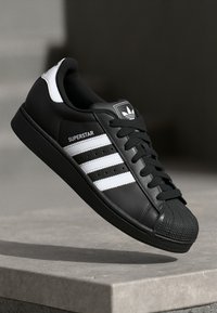 Black Adidas Superstar sneaker with white stripes and logo, floating above a light-colored concrete surface in a minimalist setting.