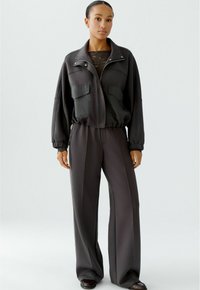 Schwarze Sportjacke mit Reißverschluss, zwei Fronttaschen und elastischen Bündchen, kombiniert mit passenden weit geschnittenen Hosen. Glatte Stoffstruktur.