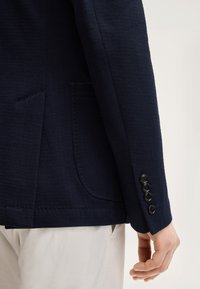 Rękaw teksturowanego granatowego żakietu i detal kieszeni z czterema czarnymi guzikami na mankiecie, noszony na jasnobeżowych spodniach, ręka swobodnie opuszczona wzdłuż ciała.