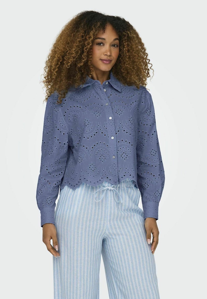 Blusa azzurro chiaro in pizzo con orlo a scallop e colletto a revers, caratterizzata da motivi a intaglio e chiusura con bottoni, abbinata a pantaloni rilassati a righe.