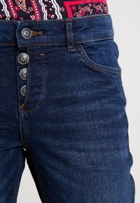 Tmavě modré džíny z denimu s vysokým pasem, pěti knoflíky v pase a jemnými prošívacími detaily. Hladká textura s mírným vyblednutím.