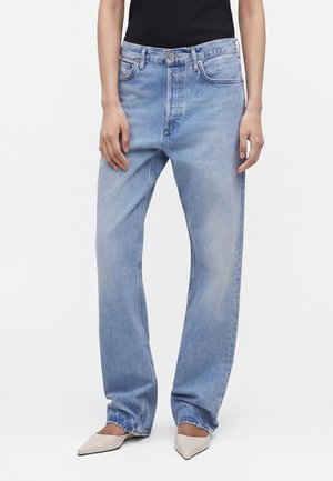 Person iført lyseblå jeans med lige ben, sort top og hvide spidse sko, stående mod en ensfarvet baggrund.
