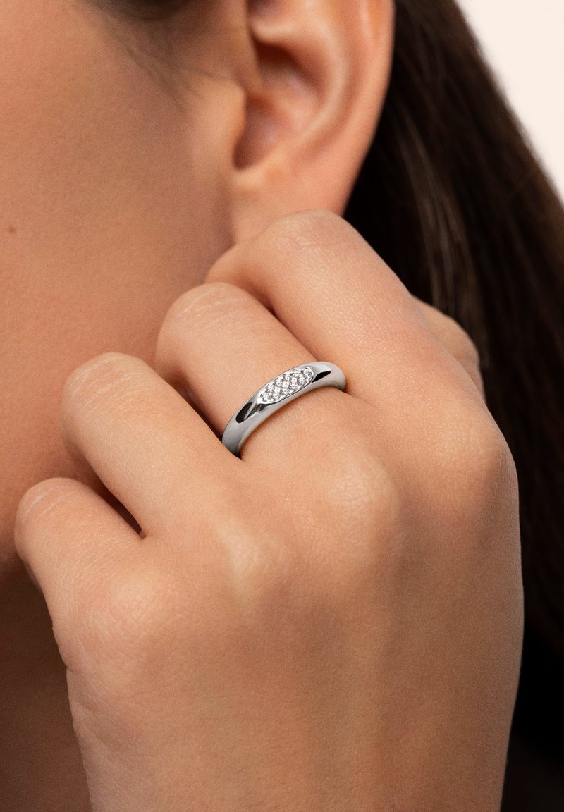 Anillo de plata con una banda lisa y una sección central que presenta pequeñas piedras claras incrustadas, llevado en una mano contra un fondo neutro.