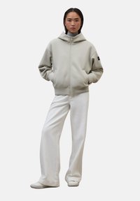 Joven de pie con las manos en los bolsillos, vistiendo una sudadera con cremallera gris claro, pantalones amplios blancos y zapatillas blancas, frente a un fondo liso.