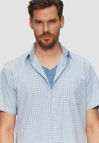 Koszula w kratę w jasnoniebieskim i białym kolorze z krótkimi rękawami, z kołnierzykiem typu spread i kieszonką z przodu. Pod nią niebieski t-shirt z dekoltem w serek.