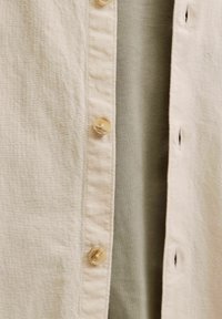 Bēša pogām aizdaļāma krekla ar teksturētu audumu, kurai ir caurspīdīgas, apaļas pogas, valkāta pāri maigi zaļai drēbei.