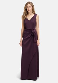 Długa, bezrękawna sukienka w kształcie litery V w głębokim fiolecie, z drapowanym przodem i akcentem w postaci kokardy z boku. Wykonana z gładkiego, lejącego się materiału.
