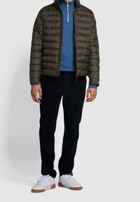 Doudoune vert olive avec coutures horizontales, col zippé, chemise à manches longues bleue en dessous, pantalon sombre et baskets blanches avec semelles beiges.