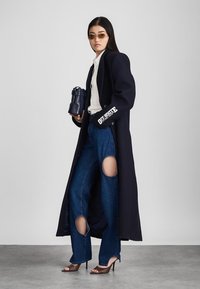 Marineblauwe lange jas, witte blouse, versleten blauwe jeans met uitsnijdingen, en open tenen hoge hakken. Donkere handtas met zilveren accenten.