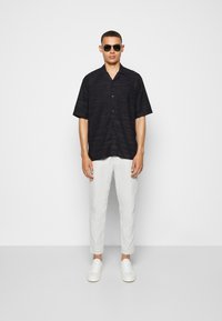 Black short-sleeve button-up shirt with a textured pattern, paired with white slim-fit trousers and white sneakers. Model wearing sunglasses.