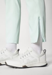 Λευκά αθλητικά παπούτσια με γκρίζες λεπτομέρειες, συνδυασμένα με ανοιχτό μυρωδένιο πράσινα παντελόνια που στενεύουν προς τα κάτω και διαθέτουν φερμουάρ στους αστραγάλους, καθώς και ριγέ λευκές κάλτσες.