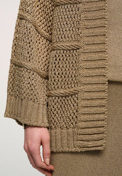 Cardigan lavorato a maglia beige con un design testurizzato, caratterizzato da sezioni in mesh aperto e polsini a coste. Mostra una mano che tiene il bordo.