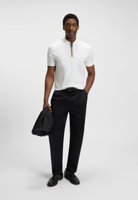 Weißes Poloshirt mit kurzen Ärmeln und Reißverschluss sowie mehrfarbigen Streifenakzenten, kombiniert mit schwarzen Hosen und schwarzen Schuhen, hält eine schwarze Jacke.