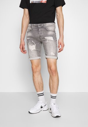 Man draagt grijze gescheurde denim shorts, zwart T-shirt, witte sokken met zwarte strepen en witte Nike sneakers, staand tegen een effen achtergrond.