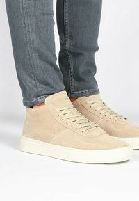 Zapatillas altas de ante beige con suelas de goma crema, que presentan un diseño de cordones y acentos en contraste en marrón claro en el tobillo.