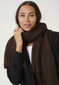 Écharpe en tricot marron à texture côtelée, drapée autour du cou, complétée par un blazer noir, avec une main reposant doucement sur le tissu.