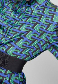 Långärmad skjorta i svart med ett djärvt grönt och blått geometriskt mönster. Har en figurnära svart midja och knäppa.