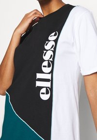 T-shirt à blocs de couleurs noir, blanc et bleu sarcelle avec un design courbé ; présente un grand logo "ellesse" en blanc sur le devant. Matière en coton.