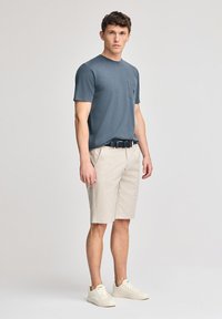 Camisa de algodón gris de manga corta para hombre con un bolsillo en el pecho, combinada con pantalones cortos beige y zapatillas deportivas blancas, de pie frente a un fondo neutro.