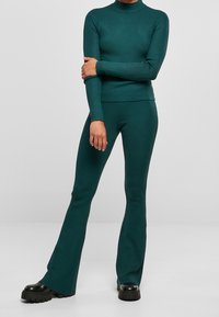 Person trägt einen tealfarbenen, gerippten Rollkragenpullover und passende ausgestellte Hosen mit schwarzen, klobigen Plateau-Schuhen und steht vor einfachem Hintergrund.