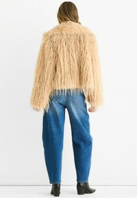 Faux fur jacket in light beige with long, shaggy texture. Features wide sleeves and a cropped fit, paired with high-waisted blue jeans.