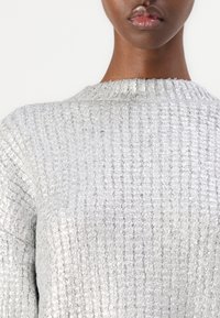 Ljusgrå, texturerad tröja med ribbat mönster. Rund halsringning och långa ärmar. Materialet verkar mjukt och något reflexivt.