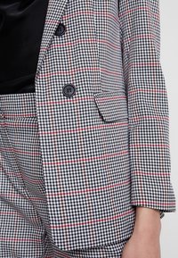 Ruudullinen bleiseri mustassa, valkoisessa ja punaisessa. Varustettu kaksirivisellä etuosalla, kahdella napilla ja pienellä taskulla vasemmalla puolella.