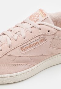 Zapatillas de ante rosa claro con una superficie texturizada, logo de Reebok en color rosa dorado y un cuello acolchado. Suela de goma blanca con detalles en forma de puntos.