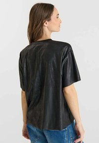 Donna con lunghi capelli castani che indossa un top nero largo a maniche corte e pantaloni in denim blu, vista di lato con la schiena visibile.