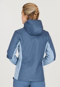 Blå jacka med en strukturerad ljusblå sidopanel, en huva och slätt tyg. Påfallande sömnadsdetaljer och figurnära design bak.