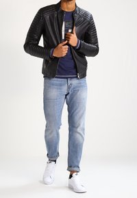 Černá kožená bunda s prošívanými rameny, spárovaná s modrou košilí, světle modrými džíny a bílými teniskami. Neformální městský outfit.