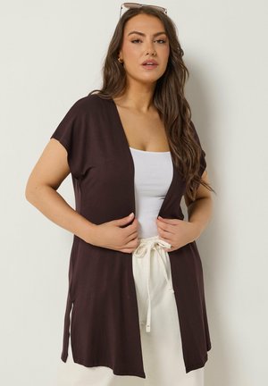 Frau mit langem Haar trägt eine dunkle offene Strickjacke über einem weißen Tanktop und cremefarbenen Hosen mit Kordelzug und richtet die Kanten der Strickjacke.