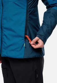 Chaqueta aislante en azul y azul oscuro con cremallera frontal, puños ajustables y un tirador naranja en un bolsillo lateral.