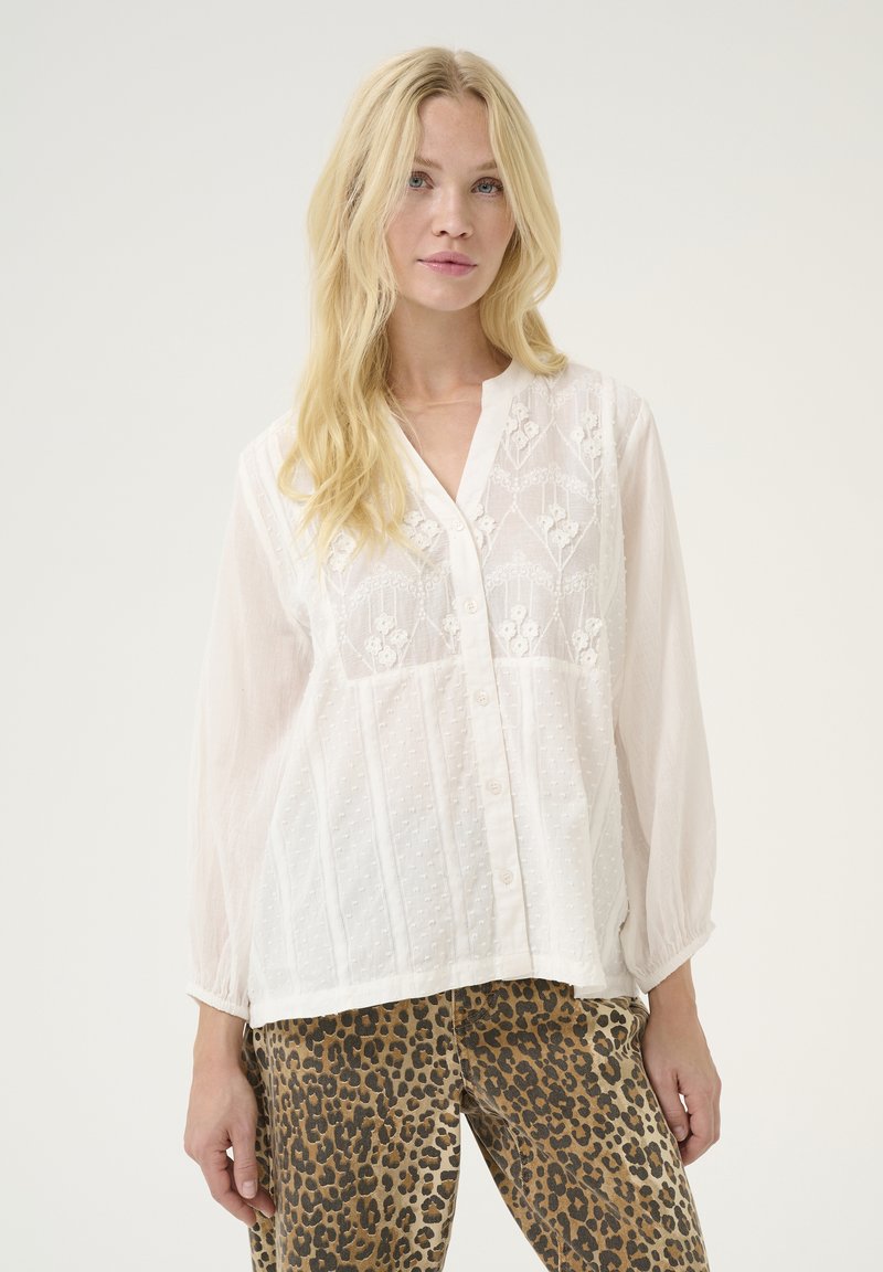 Blusa bianca, strutturata, con ricami floreali e chiusura a bottoni. Presenta maniche lunghe e leggermente a sbuffo e una vestibilità comoda.
