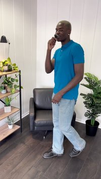 Camisa polo azul, jeans azuis claros com bainhas desfiadas e sapatos pretos. Ambiente interior com plantas e uma cadeira de couro preta.