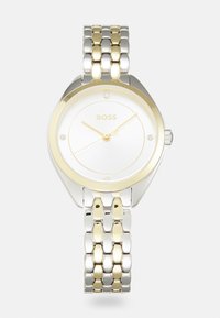 Montre en acier inoxydable argenté et doré avec un cadran rond blanc, trois aiguilles dorées et quatre petits accents en cristal. Le bracelet présente un design bicolore.