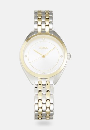 Montre en acier inoxydable argenté et doré avec un cadran rond blanc, trois aiguilles dorées et quatre petits accents en cristal. Le bracelet présente un design bicolore.