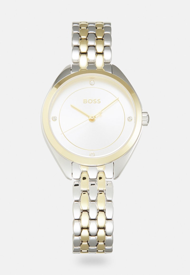 Montre en acier inoxydable argenté et doré avec un cadran rond blanc, trois aiguilles dorées et quatre petits accents en cristal. Le bracelet présente un design bicolore.