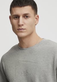 Szürke, apró rácsos pulóver, kerek nyakkivágással és hímzett "ABC" logóval a mellkasán, amelyhez ezüstlánc nyaklánc társul.