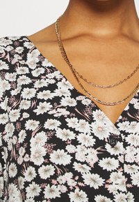 Svart tyg med blommönster i vitt och rosa, med V-ringning och knappdetalj, kompletterat av lager av gyllene kedjor.