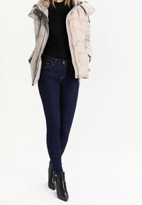 Mujer vestida con una chaqueta acolchada beige, suéter negro, jeans ajustados azul oscuro y botas negras con tobillo, de pie con las manos en los bolsillos.