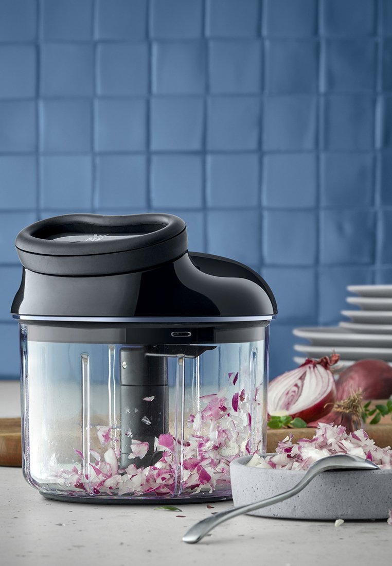 Elektrischer Zerkleinerer mit schwarzem Deckel und klarer Glasschüssel, gefüllt mit fein gehackten roten Zwiebeln, auf einer hellen Oberfläche mit daneben stehenden Tellern.